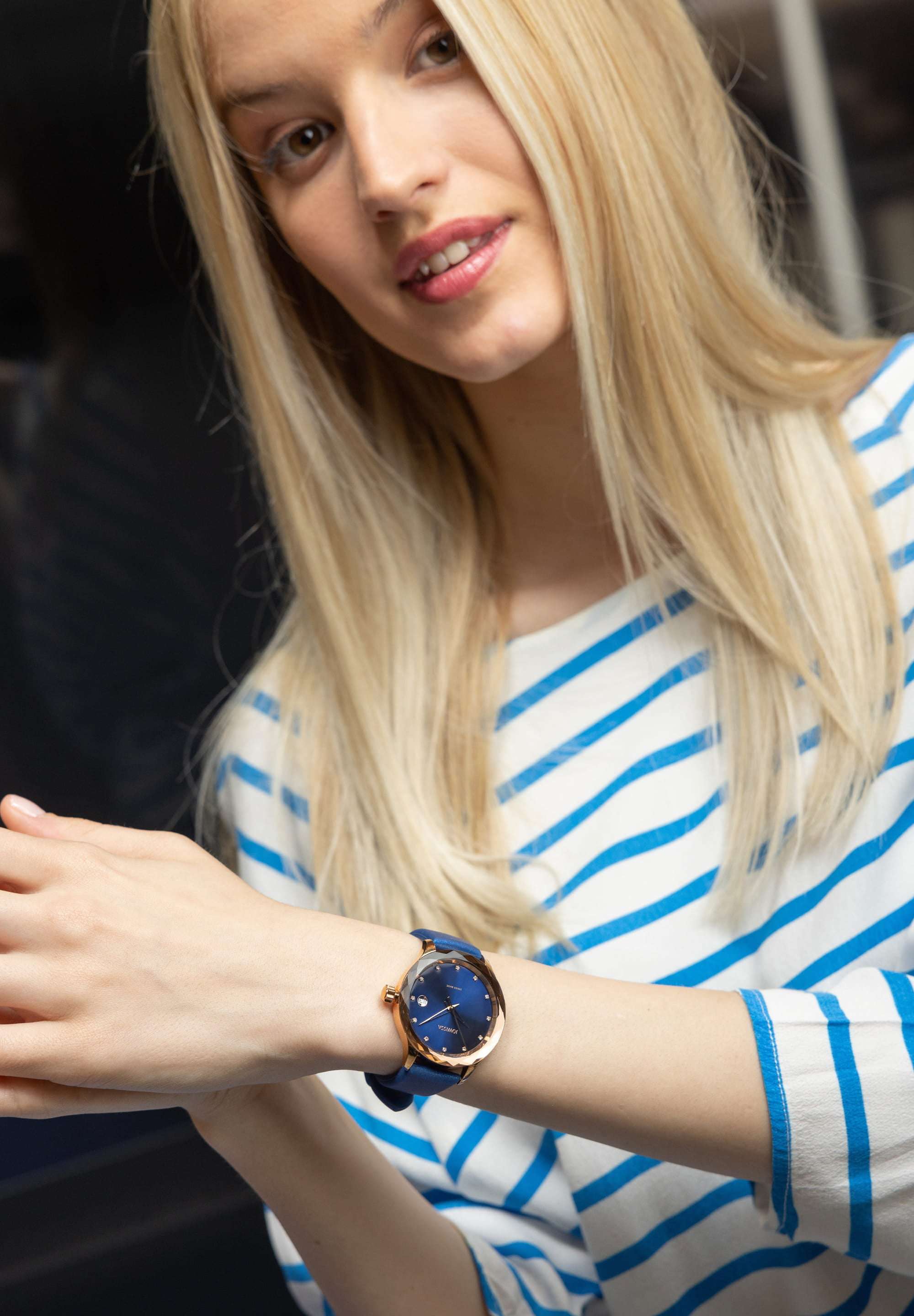 blue watch worn by a blonde girl with white t shirt - the girl is looking at the camera showing the watch - the watch is made in switzerland - the watch has a stainless steel case