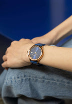 Blue watch made for women - worn by a girl sitting on a chair on a blue background - the watch is all made in switzerland - the girl has placed the hand on her knee 
