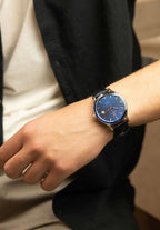 Blue mens watch with black leather strap - The watch is designed for comfort and durability, making it perfect for everyday wear or special occasions - worn by a men who is wearing white t-shirt blue shirt and grey pants 