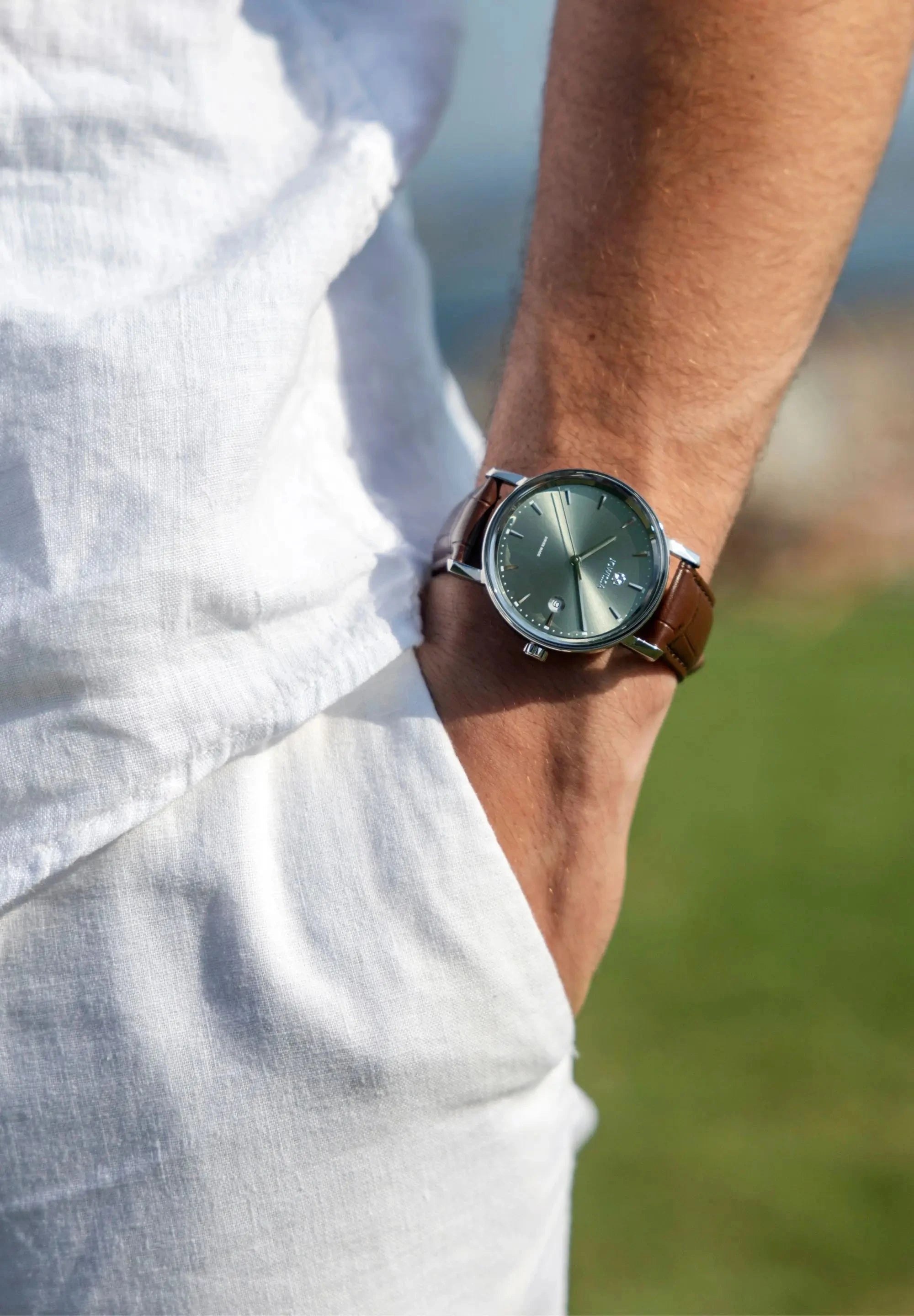 Swiss men watch - with brown leather strap and green dial with silver details - worn by a man with white pants and shirt - all swiss made