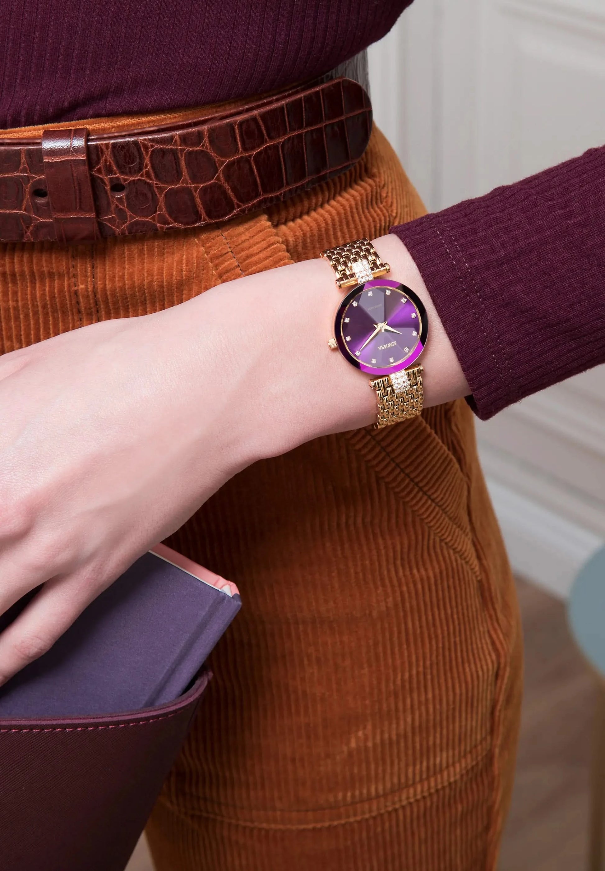 Woman wearing a gold watch with a purple face, paired with brown pants and a maroon top. The watch is Swiss Made by Jowissa, the collection is Facet Strass.