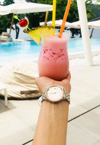 J4.229.M  medium size watch - worn by a woman with a glass of pina colada on her hand – features a stainless steel case – boasts a shiny silver dial with rose-gold hour markers – includes a calendar window - all made in switzerland