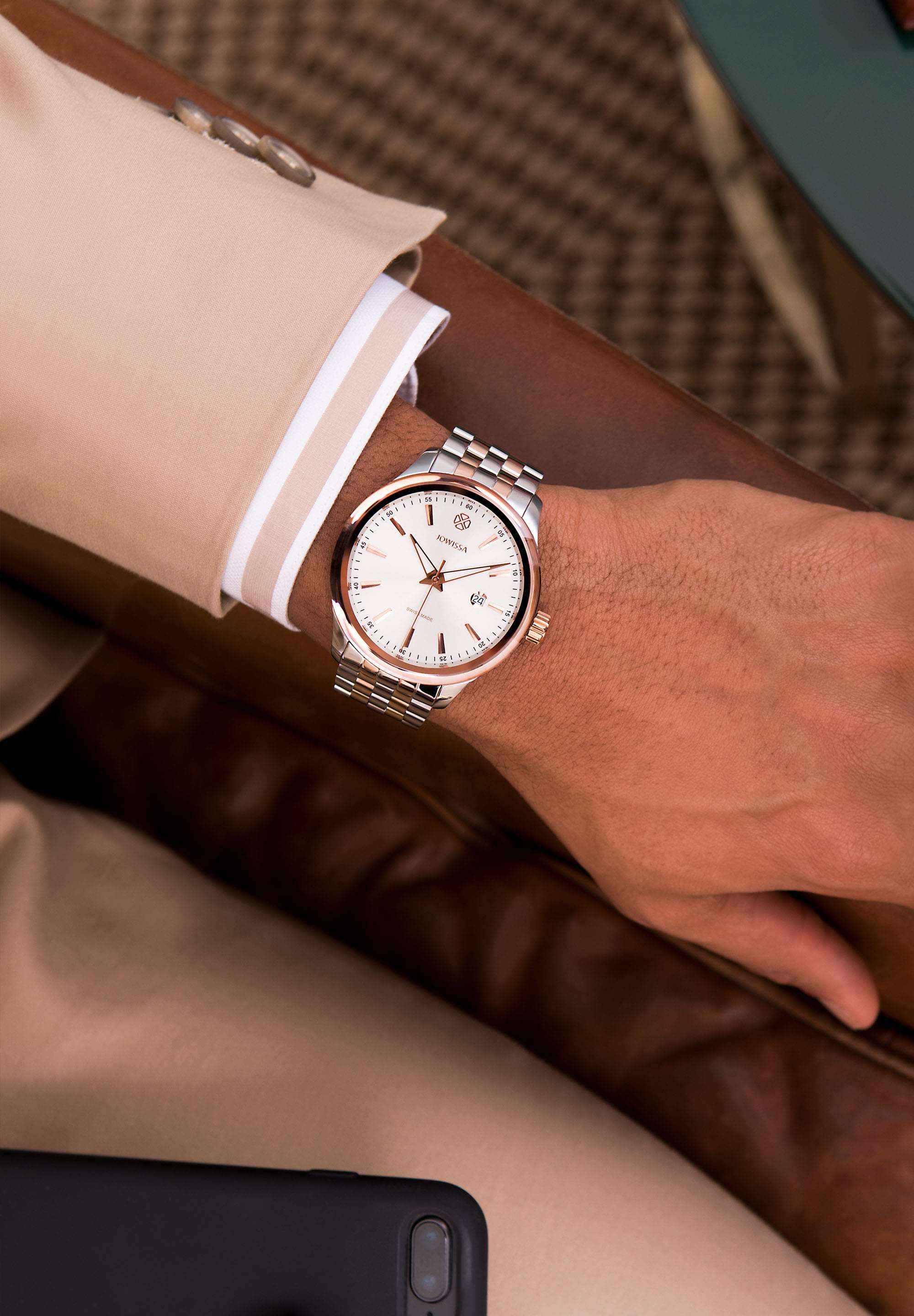 Tiro Swiss Men's Watch J4.229.L with a bi-colour stainless steel bracelet, 45mm case, and silver dial featuring rose-gold hour markers- worn by a man that is seated on a brown couch.