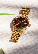 Gold womens watch with red details  features a gold-plated stainless steel case with a glossy polished surface and a warm brown sunray dial. With a date window, 5 ATM water resistance, and a diamond-cut bezel