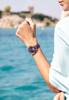 J6.240.M Swiss Watch – This elegant women’s watch features a rose-gold stainless steel case paired with a vibrant purple sunray dial, adorned with 12 rhinestone hour markers for a touch of glamour - it includes a practical date window. The genuine patent leather strap offers both comfort and style, while the 5 ATM water resistance ensures durability.  Worn by a girl who is posing in front of the beach wearing a blue shirt