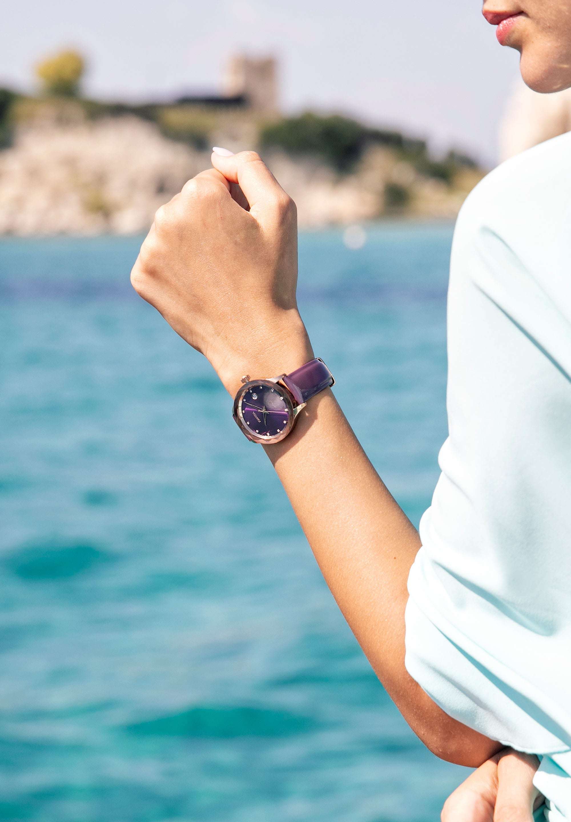 J6.240.M Swiss Watch – This elegant women’s watch features a rose-gold stainless steel case paired with a vibrant purple sunray dial, adorned with 12 rhinestone hour markers for a touch of glamour - it includes a practical date window. The genuine patent leather strap offers both comfort and style, while the 5 ATM water resistance ensures durability.  Worn by a girl who is posing in front of the beach wearing a blue shirt