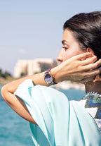 J6.240.M Watch for women –  with a rose-gold stainless steel case and a  purple sunray dial, accented by 12 sparkling rhinestones as hour markers. Made in switzerland - worn by a girl who is in front of the beach looking aside